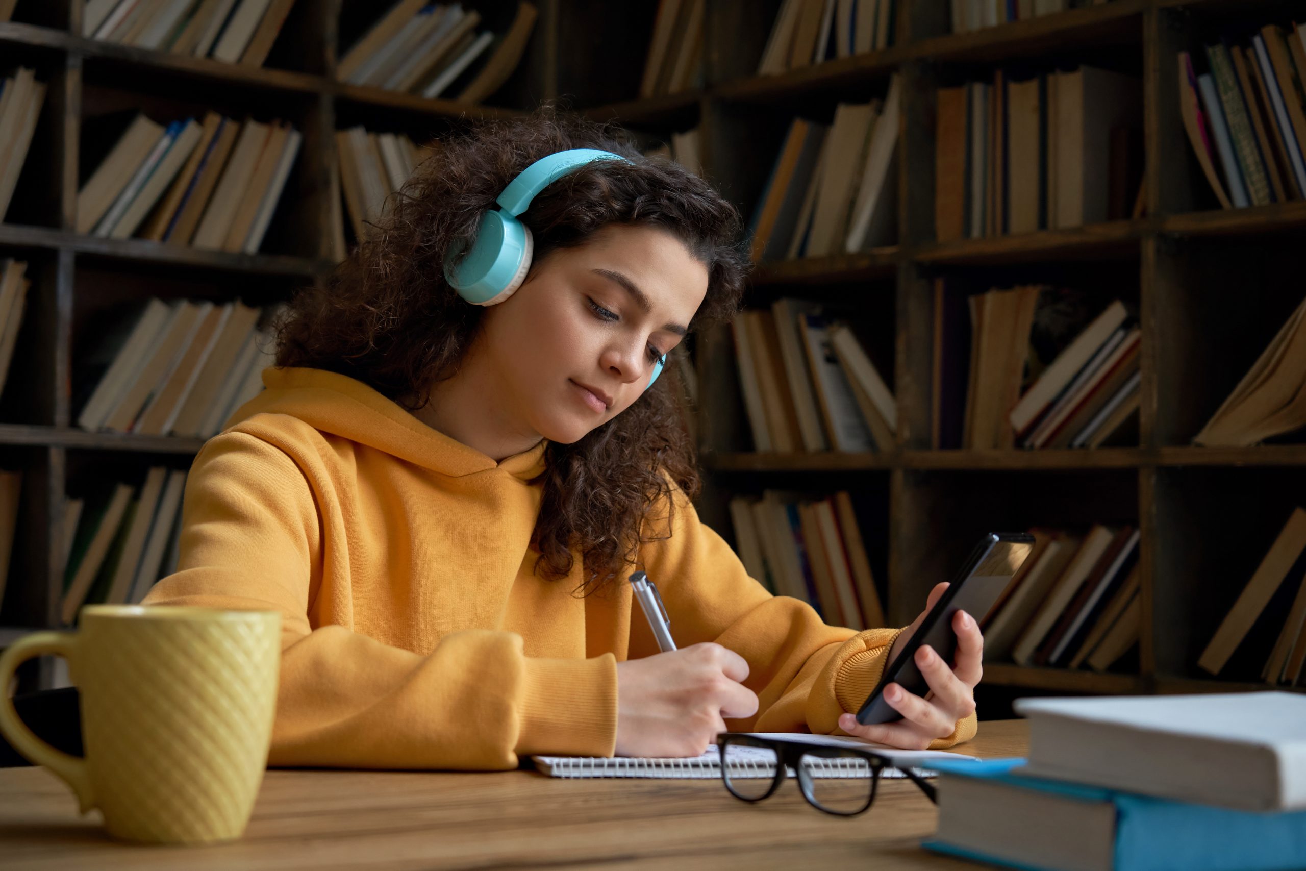 Student wearing headset and holding a smart phone while writing in a notebook in a library, with a mug and glasses in the background.