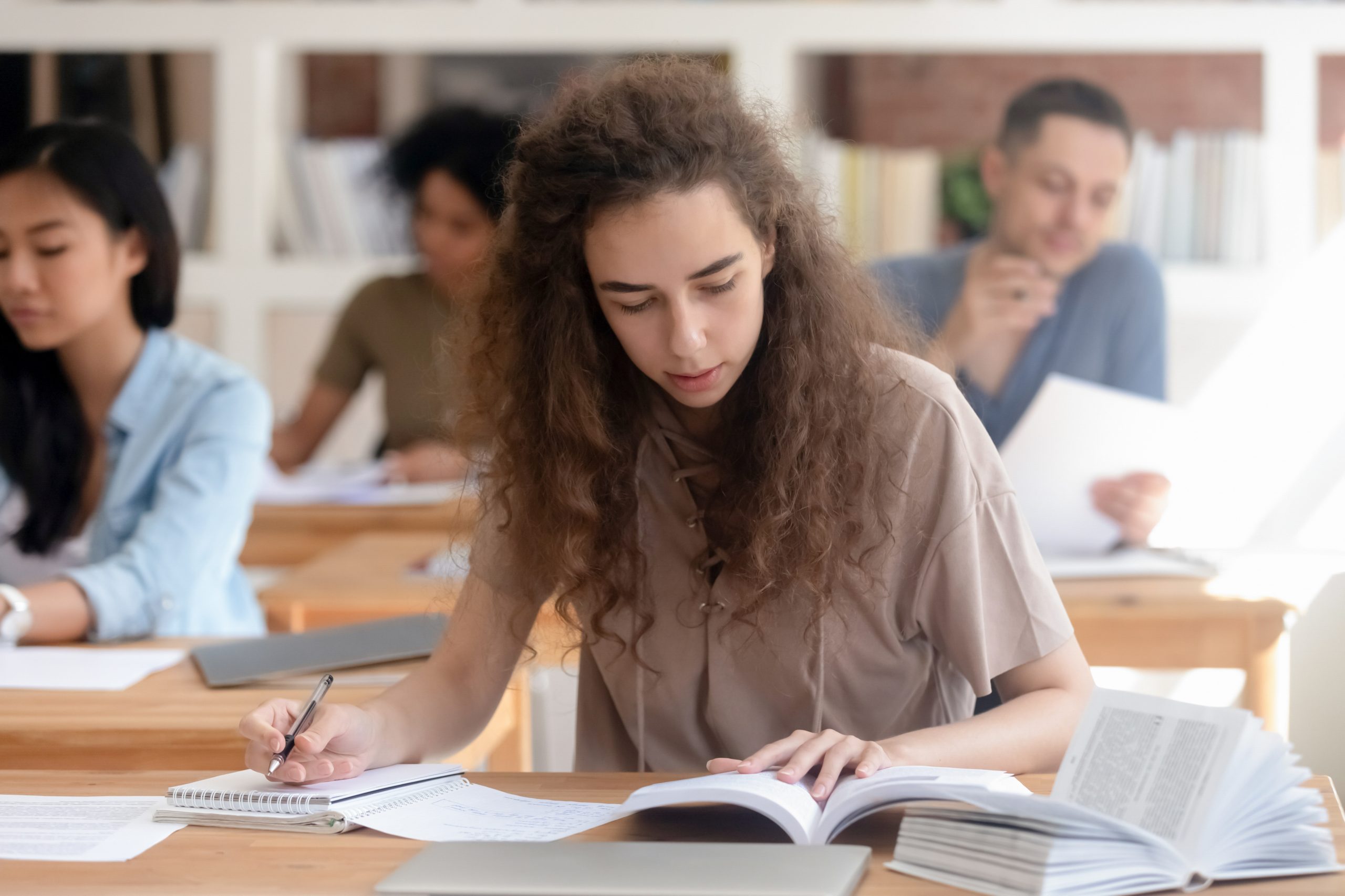 Student reading textbook and writing with other students in background